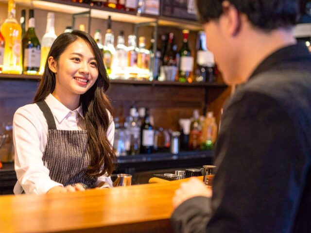 バーカウンター越しに笑顔で接客するエプロン姿の女性バーテンダー