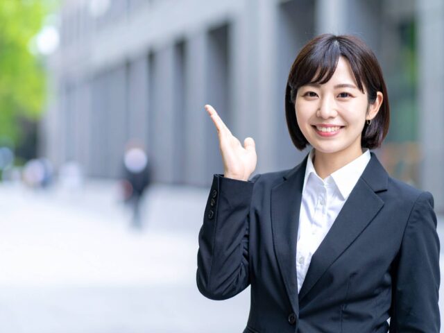 オフィス街を背景に笑顔で案内するスーツ姿の日本人女性
