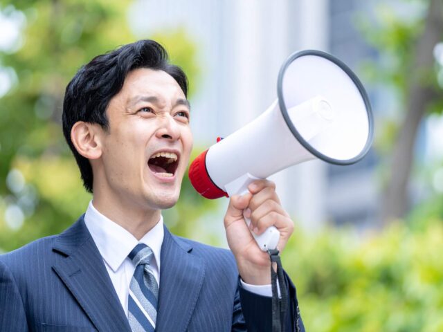 屋外でメガホンを使って大声で叫ぶスーツ姿の日本人男性