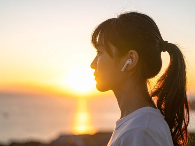 夕日を背景にワイヤレスイヤホンで音楽を聴く女性の横顔シルエット