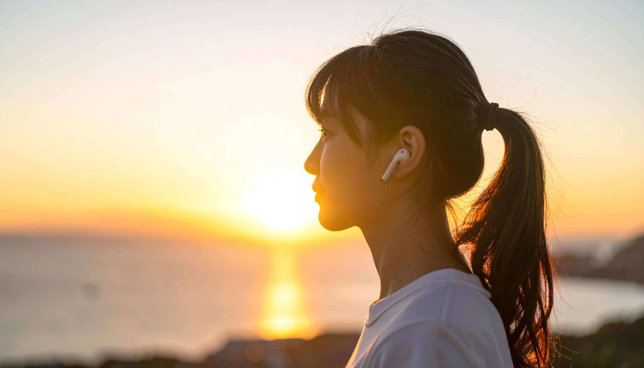夕日を背景にワイヤレスイヤホンで音楽を聴く女性の横顔シルエット